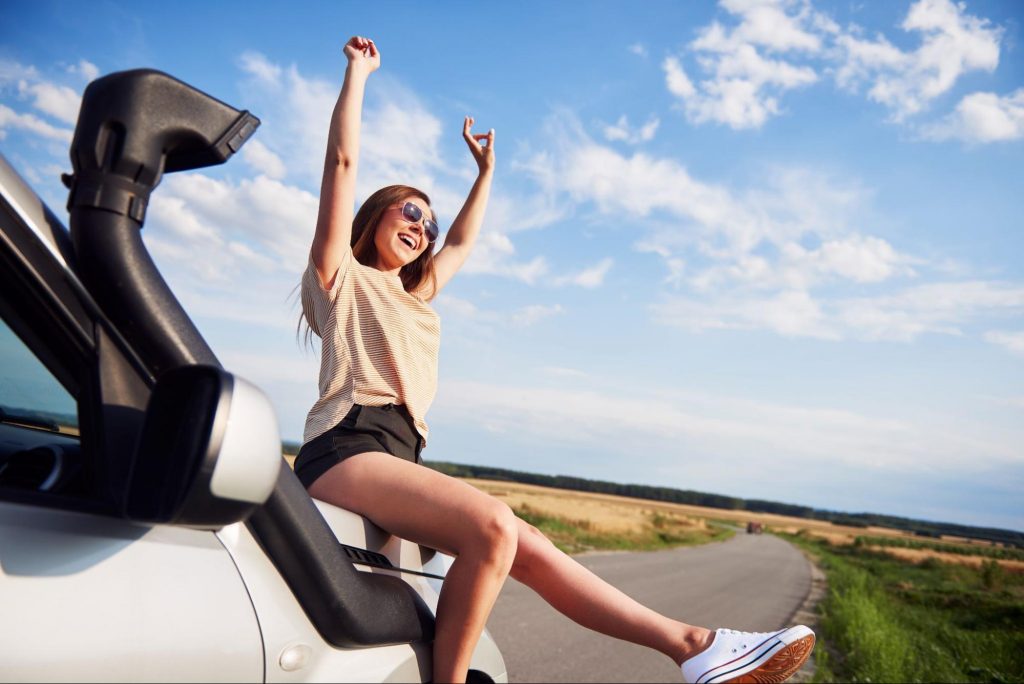 A happy woman sitting on a car in the middle of a country road