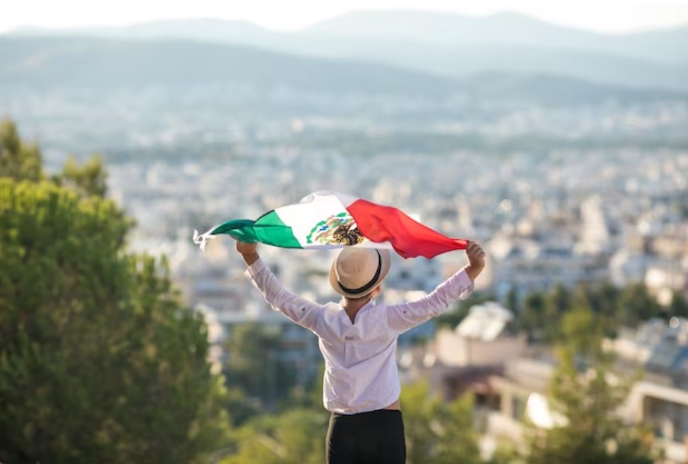 a person wearing a hat and a white shirt while holding a flag of Mexico and looking at the city landscape from a distance