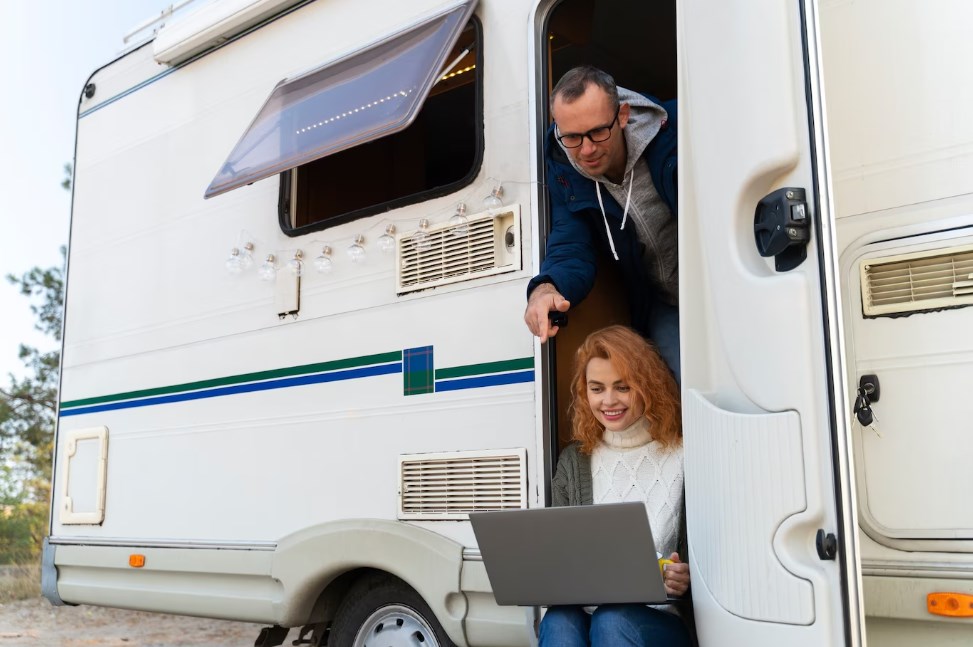 a couple in the doorway of a white RV trailer with a laptop